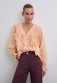 Mujer vistiendo una blusa de melocotón con volantes y un sutil estampado de leopardo, metida en unos pantalones oscuros púrpuras de cintura alta, de pie contra un fondo sencillo.