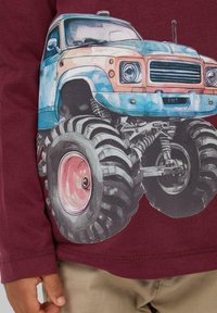 Bordeauxfarbenes Langarmshirt mit einem bunten Grafikdesign eines Trucks mit übergroßen schwarzen Reifen und rosa Felgen, das komplexe Details und Farbnuancen hervorhebt.