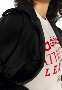 Zwarte bomberjack met geribde kraag en rits, gedragen over een wit t-shirt. Rode tekst en logo op het t-shirt zijn duidelijk zichtbaar.