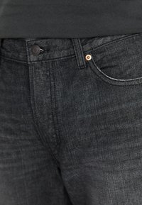 Jeans de mezclilla negra con textura desgastada, herrajes de cobre en la cintura y sutilezas en los detalles de las costuras en los bolsillos frontales.