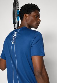 Blauw sportshirt van gestructureerde stof, met een wit verticaal logo op de achterkant. Een tennisracket rust op de schouder.
