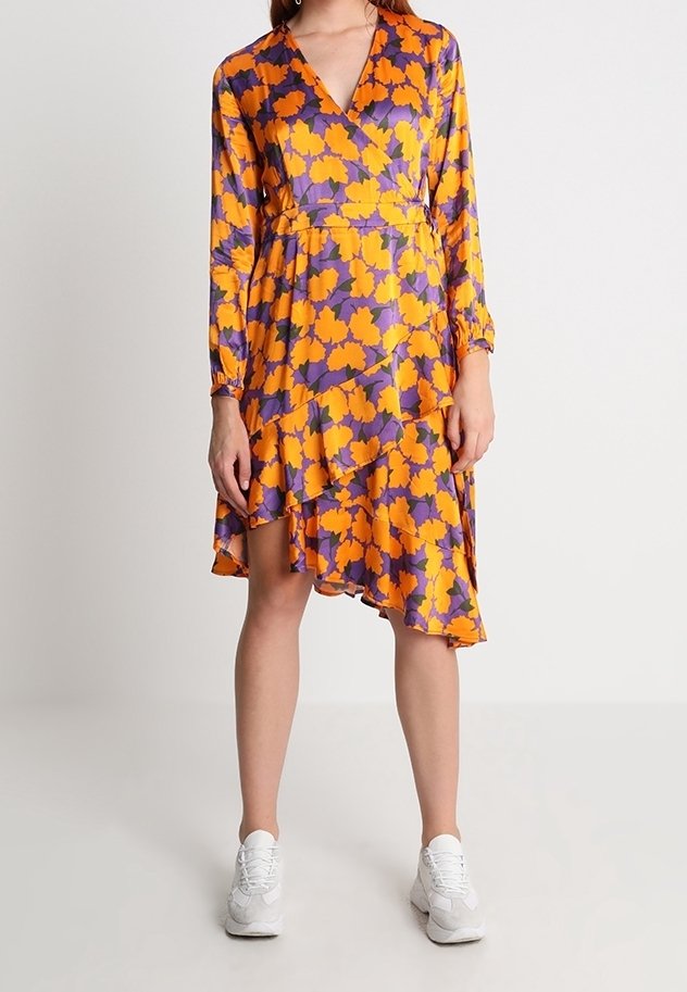Femme portant une robe longue à manches longues, col en V, asymétrique, à fleurs orange et violettes, associée à des baskets blanches, debout devant un fond uni.