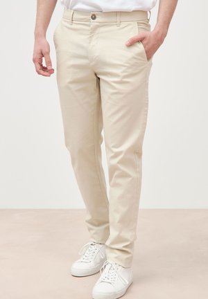 Mann trägt beige Chino-Hosen mit Knopf und Reißverschluss, eine Hand in der Tasche, kombiniert mit weißen Turnschuhen auf einem neutralen Boden.
