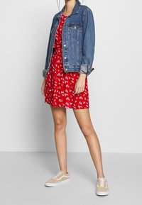 Persona che indossa un vestito floreale rosso, una giacca di jeans blu con le maniche arrotolate e scarpe da ginnastica beige, in piedi contro uno sfondo chiaro e uniforme.