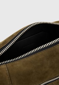Interior de un bolso de ante verde oliva con un forro negro, que cuenta con un cierre de cremallera plateado y un pequeño tarjetero negro visible en su interior.