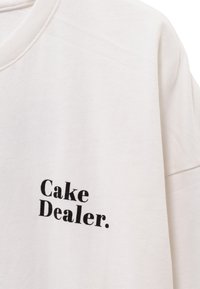 Camiseta de algodón blanca con cuello redondo, que presenta "Cake Dealer" impreso en negrita con texto negro en el frente. Textura suave, diseño casual.