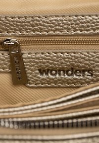 Pochette dorée métallisée avec une finition texturée, dotée d'une tirette de fermeture éclair gravée du mot "merveilles", placée sur un fond en tissu beige.