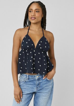 Jeune femme aux cheveux tressés, portant un haut dos nu bleu marine à motifs floraux blancs et un jean bleu clair, debout avec une main dans la poche.