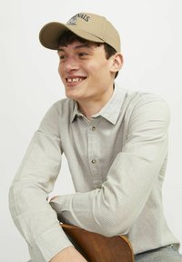 Cappellino da baseball beige con logo, camicia a righe grigia con bottoni, seduto con le braccia conserte su una sedia di legno, sfondo neutro.