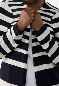 Krémový a námořnicky modrý pruhovaný cardigan s dlouhými rukávy, otevřeným předním dílem a texturovanou pletenou tkaninou. Zaměření na ruce držící oděv.