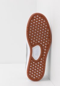 Semelle en caoutchouc d'une basket Vans, présentant une semelle extérieure marron texturée avec des motifs en forme de losange, une tige en daim gris et une bordure en caoutchouc blanche.