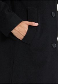 Cappotto di lana nera con tasca laterale, chiusura a bottoni e una finitura morbida e testurizzata. Linee pulite con dettagli minimali.