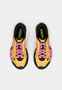 Une paire de chaussures de course jaunes et noires avec des lacets roses et des semelles blanches, vue de dessus sur un fond blanc uni.