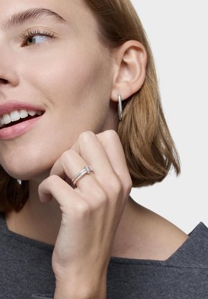 Mujer con cabello corto castaño claro, lleva pendientes de aro plateados y un anillo plateado, sonriente y tocándose ligeramente la barbilla.