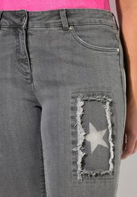 Jeans de mezclilla gris con un parche desgastado que tiene una estrella blanca. Incluyen un diseño tradicional de cinco bolsillos y bordes deshilachados.