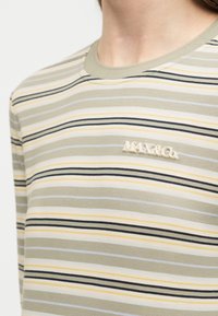 Άτομο που φοράει μακρυμάνικο πουκάμισο με ρίγες σε μπεζ, μαύρο και ανοιχτό κίτρινο, με κεντημένο το λογότυπο "MAX&Co." στο στήθος.