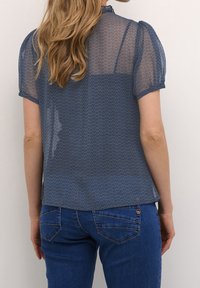 Blå genomskinlig blus med korta puffärmar och hög krage, med ett geometriskt mönster och en slät, lätt struktur.