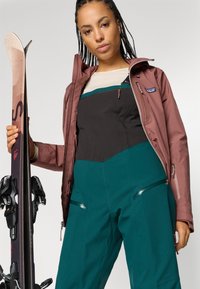 Εξοπλισμός σκι με ένα μπορντό μπουφάν, μαύρη μπλούζα και τιρκουάζ σαλοπέτα με τσέπες που κλείνουν με φερμουάρ, κρατώντας ένα σκ�ι και τις κράνες με το άλλο χέρι.