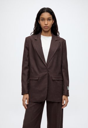 Une femme porte un tailleur marron foncé à fines rayures, composé d’un blazer oversize et d’un pantalon assorti sur une chemise blanche, devant un fond uni.
