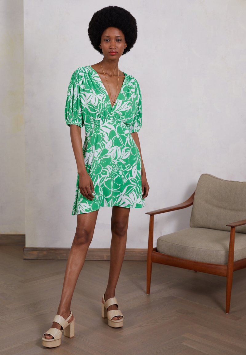 Vestido floral verde com mangas bufantes e decote em V profundo. Apresenta uma cintura franzida e está combinado com sandálias de plataforma bege. Piso de madeira texturizada.