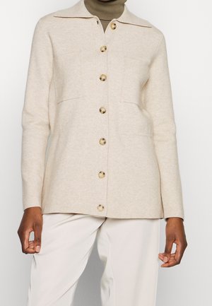 Cardigan beige en laine à boutons avec un col, deux poches avant et de gros boutons ronds, porté par-dessus un col roulé beige. Texture douce.