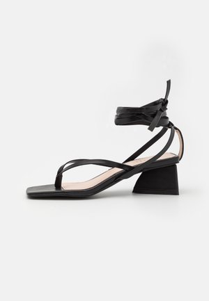 Sort sandal med firkantet tå, tynd snøre-design og hæl bagpå. Har en indpakket ankelrem for sikker pasform. Glat tekstur.