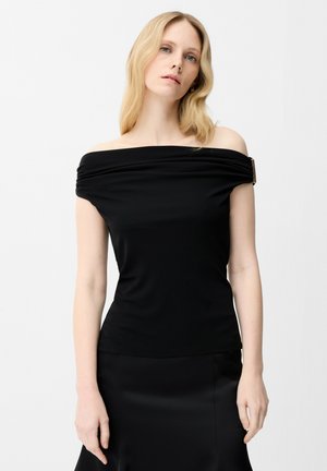 Kobieta o blond włosach, ubrana w czarny top z odkrytymi ramionami z detalem w postaci klamry oraz dopasowaną czarną spódnicę, stojąca na jednolitym tle.