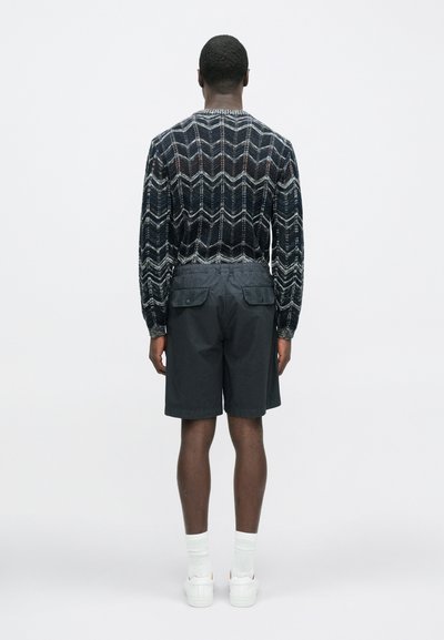 Sweter dziergany z geometrycznym wzorem w ciemnych kolorach, połączony z ciemnymi szortami typu cargo z dwoma tylnymi kieszeniami, białymi skarpetkami i trampkami.