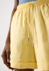 Yellow linen shorts with an elastic waistband and a braided drawstring. Features side pockets and a rolled hem. Smooth texture.