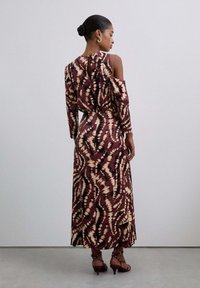 Mujer vestida con un largo vestido de color marrón y crema con un patrón, con un hombro al descubierto, de pie de espaldas a la cámara frente a un fondo liso.