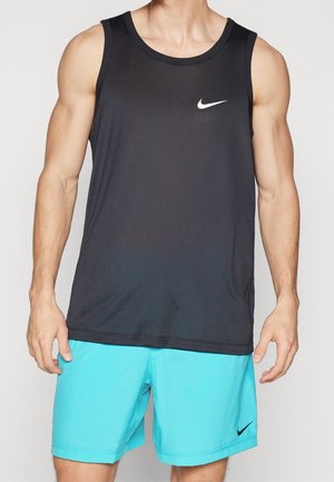 Mężczyzna ubrany w czarną, bezrękawnikową sportową koszulkę Nike i jaskrawo niebieskie krótkie spodenki Nike, stojący na jasnym tle.