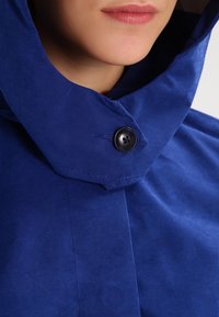 Blå huva jacka med texturerad tyg. Inkluderar en närbild av en svart knapp och en vinkelrad krage design.