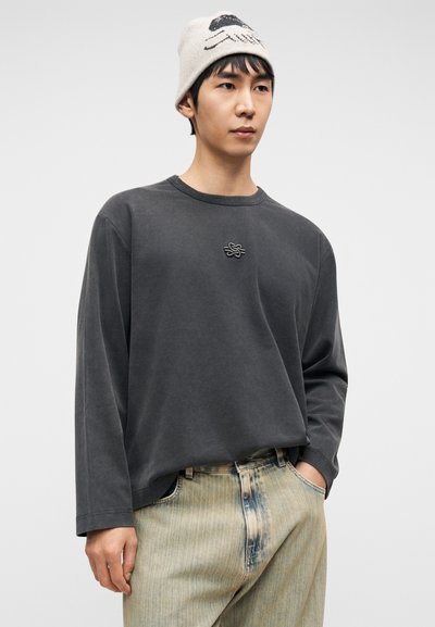 Γκρι μακρυμάνικη μπλούζα με στρογγυλή λαιμόκοψη και κεντημένο λογότυπο, συνδυασμένη με τζιν με ανοιχτό ξέβαμμα και κάθετες ρίγες, και σκουφάκι.