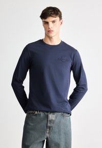 Mornarsko dolg rokav majica iz bombaža s krožnim izrezom, ki vsebuje diskretno embosirano logotip na levem prsnem delu in sproščen kroj. V kombinaciji s svetlo modrimi kavbojkami.