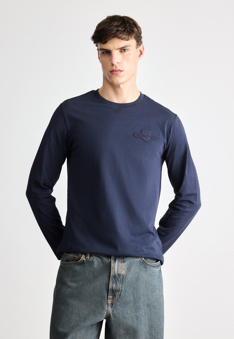 Tricou lung cu mânecă, de culoare bleu marin, confecționat din bumbac, cu guler rotund, având un logo discret embossat pe pieptul stâng și o croială lejeră. Asortat cu blugi de culoare albastru deschis.