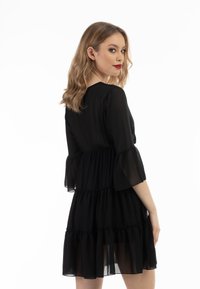 Rochie din chiffon negru cu mâneci trei sferturi tip clopot, talie strânsă, volane suprapuse și o siluetă lejeră, fluidă.
