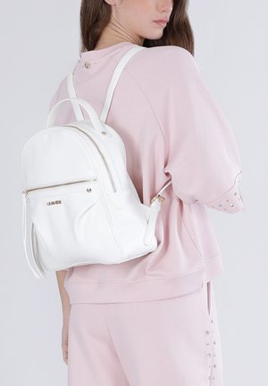 Žena v lehce růžovém neformálním outfitu, která má na zádech bílý kožený batoh se zlatým zipem a logem.