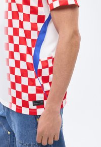 Άτομο που φοράει αθλητική φανέλα με κόκκινο και άσπρο καρό, μπλε πλαϊνές πινελιές και μπλε τζιν, με το χέρι χαλαρό στο πλευρό.