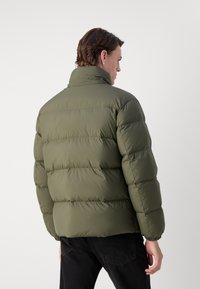 Grüne Pufferjacke mit hohem Kragen und gesteppten horizontalen Abschnitten. Der Stoff wirkt glatt und leicht glänzend. Enganliegende Bündchen.