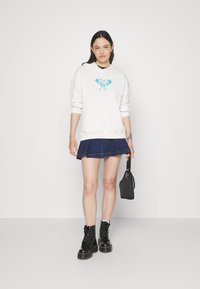 White hoodie with a blue graphic logo, paired with a dark denim skirt and black combat boots. Model holds a small black bag.
