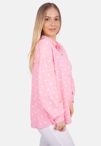 Rosa Hemd mit Knopfverschluss und weißen Herzmustern, aus strukturiertem Material. Hat lange Ärmel und eine lockere Passform, gezeigt über weißen Hosen getragen.