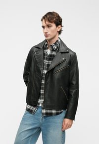 Casaco de motoqueiro em pele preta com colarinho em ponta, bolsos com fecho éclair e um corte ligeiramente cropped, sobre uma camisa xadrez em preto e branco.
