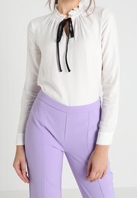 Vit texturerad blus med en volangkrage och svarta snören, ihop med ljuslavendelfärgade byxor med framseams och en kroppsnära design.