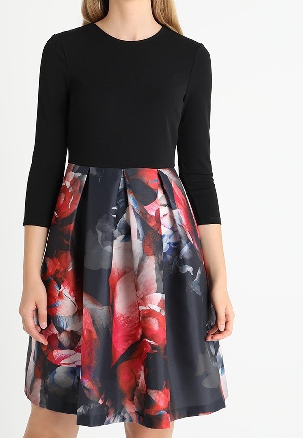 Femme portant une robe noire à manches trois-quarts avec une jupe plissée présentant un motif floral rouge, bleu et gris.