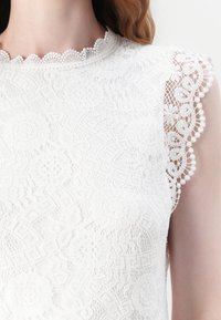 Vit spetsblus med blommönster, vågig halsringning och intrikat spetsdekoration på axlarna. Lätt och strukturerad tyg.