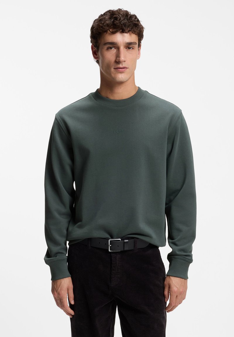 Homme portant un sweat-shirt vert foncé à col rond et un pantalon noir, debout devant un fond blanc uni.