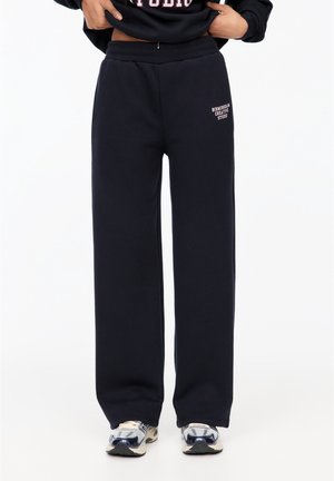 Personne portant un pantalon de jogging noir ample avec le texte « Birmingham Creative Studio » sur la cuisse et des baskets blanches, les mains dans la taille.