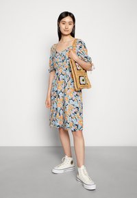 Vestido floral em azul escuro com padrões em laranja e azul claro, mangas curtas em forma de babado, até ao joelho. Acompanhado por uma bolsa trançada com detalhes coloridos.