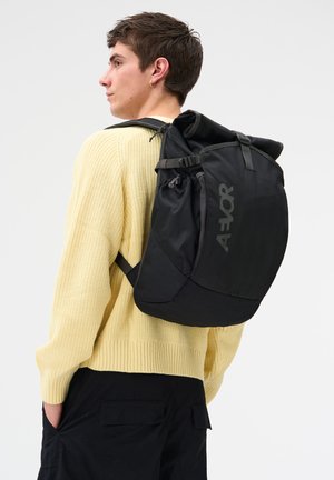 Young person with short hair wearing a yellow knit sweater and black pants, carrying a large black AER backpack over one shoulder.