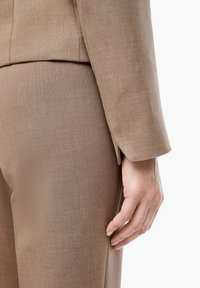 Costume marron clair sur mesure avec une finition texturée. Gros plan sur les détails de la manche et de la jambe du pantalon, mettant en valeur des lignes épurées et des éléments de design subtils.